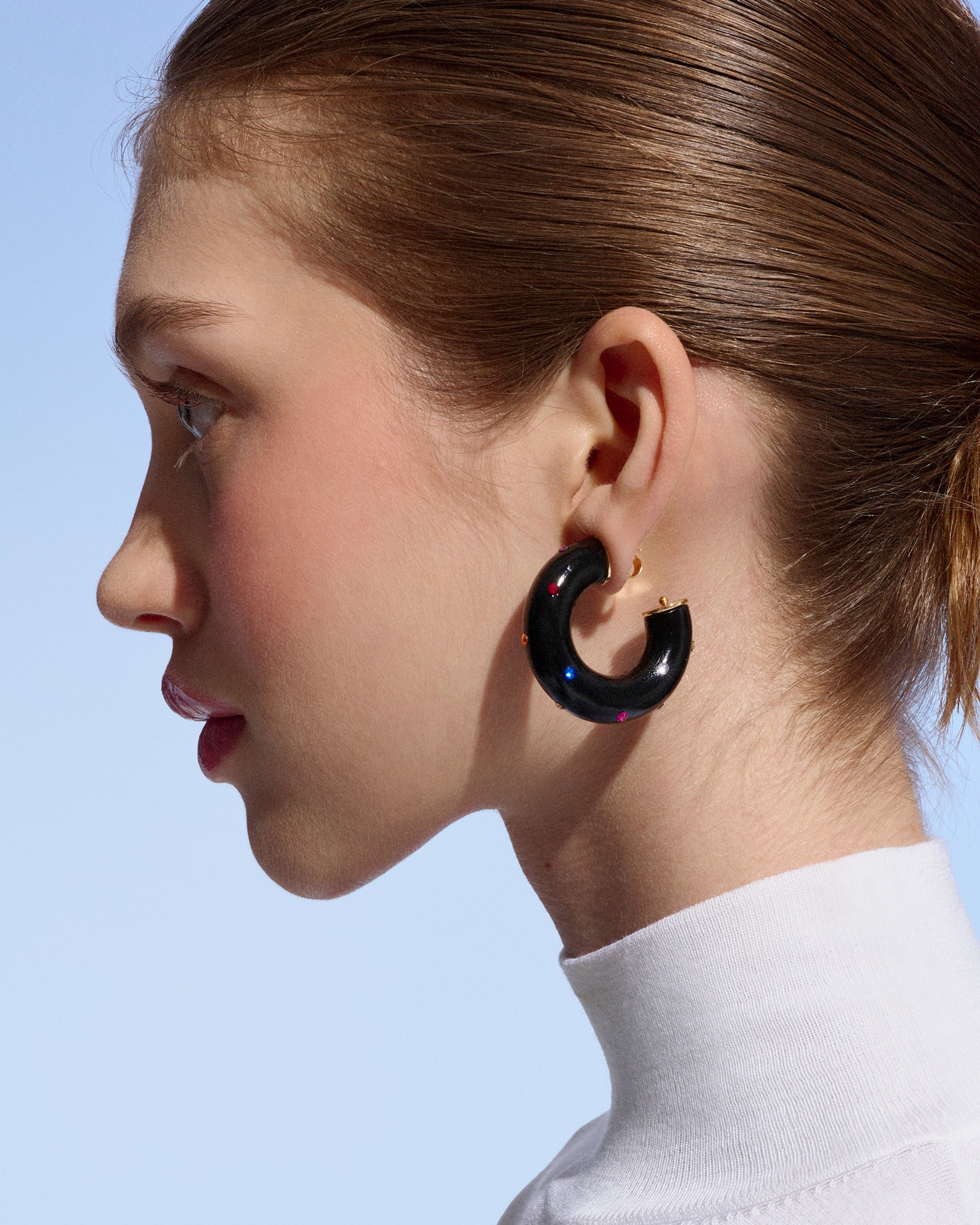 side profile of a model wearing black Alba Hoops with colorful crystals against a light blue background
